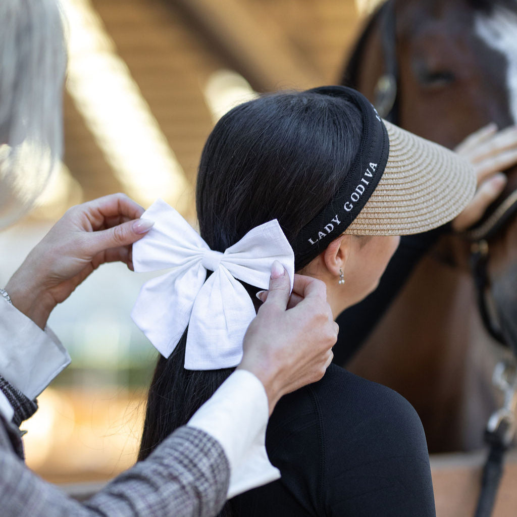 Person adjusting a white bow on another person's head with a horse in the background